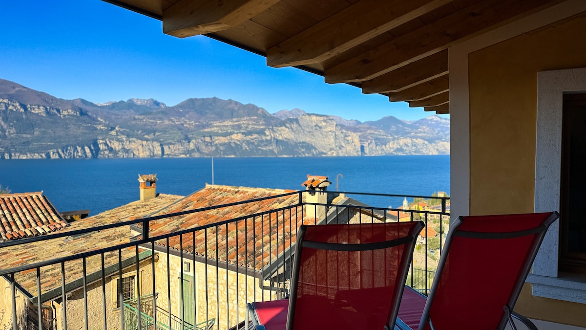A terrace with two red chairs offers a view of the serene blue lake and surrounding mountains. The rustic rooftops of nearby buildings are visible, framed by clear skies above. Natural light brightens the space, highlighting the wooden beams of the terrace overhead.