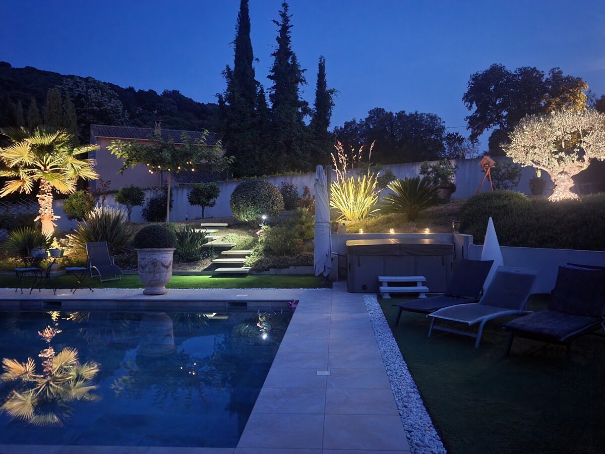 A serene outdoor space is illuminated at night, featuring a tranquil pool reflecting ambient lighting. Lush greenery and carefully arranged plants create a calming atmosphere. Comfortable loungers are positioned nearby, inviting relaxation under the starlit sky.