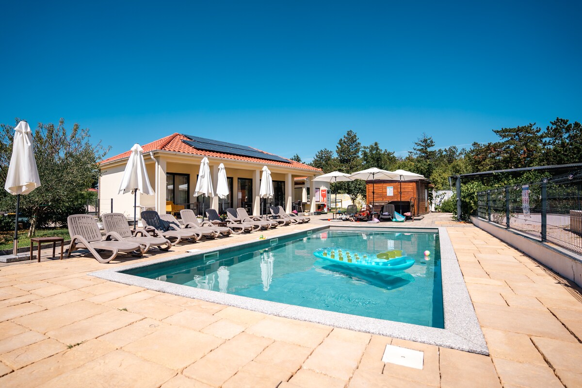 An outdoor pool is surrounded by a well-maintained patio area featuring loungers and umbrellas. A shaded seating area is visible in the background, providing a relaxing space. The clear blue sky complements the tranquil setting.
