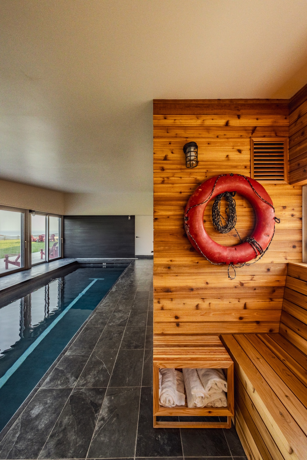 A serene indoor spa area features a swimming pool alongside a wooden paneled wall. A life preserver hangs prominently on the wall, while fresh white towels are neatly stacked on a wooden shelf. Large windows offer a view of the surrounding landscape.