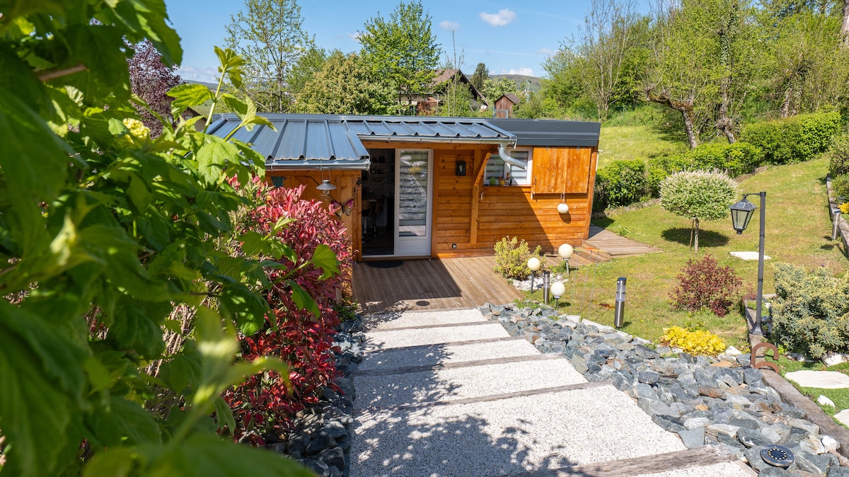 The charming chalet is framed by lush greenery and colorful landscaping, with a well-maintained pathway leading to the entrance. The exterior features wooden paneling and a sloped metal roof. Soft lighting illuminates the garden, enhancing the inviting atmosphere of the outdoor space.