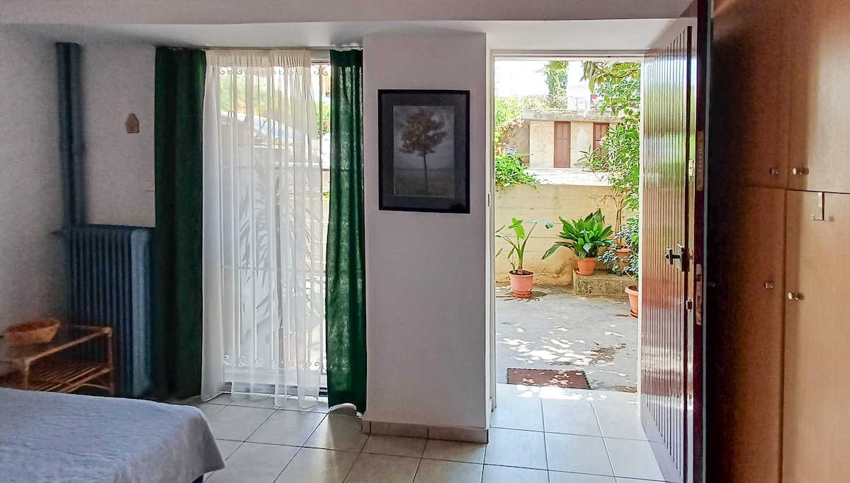 A warm interior is visible, featuring tiled flooring and natural light filtering through sheer curtains. A framed piece of art adorns the wall, while a door opens to a private yard with potted plants. Green drapes complement the space.