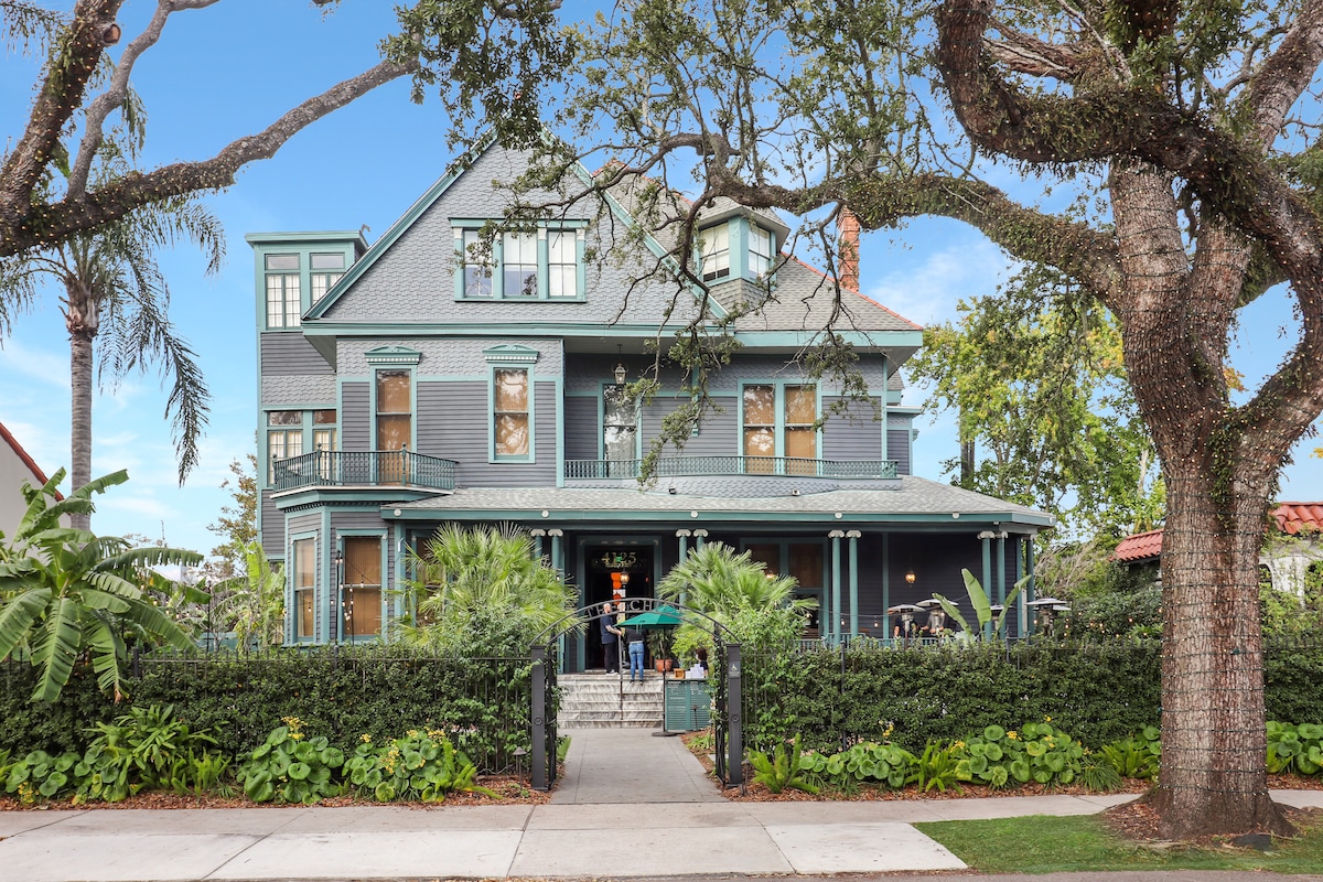 A charming two-story home features a distinctive blue exterior and multiple gabled roofs. Lush greenery surrounds the property, with palms and other plants framing the entrance, which is accessed via a set of stairs leading to a spacious front porch.