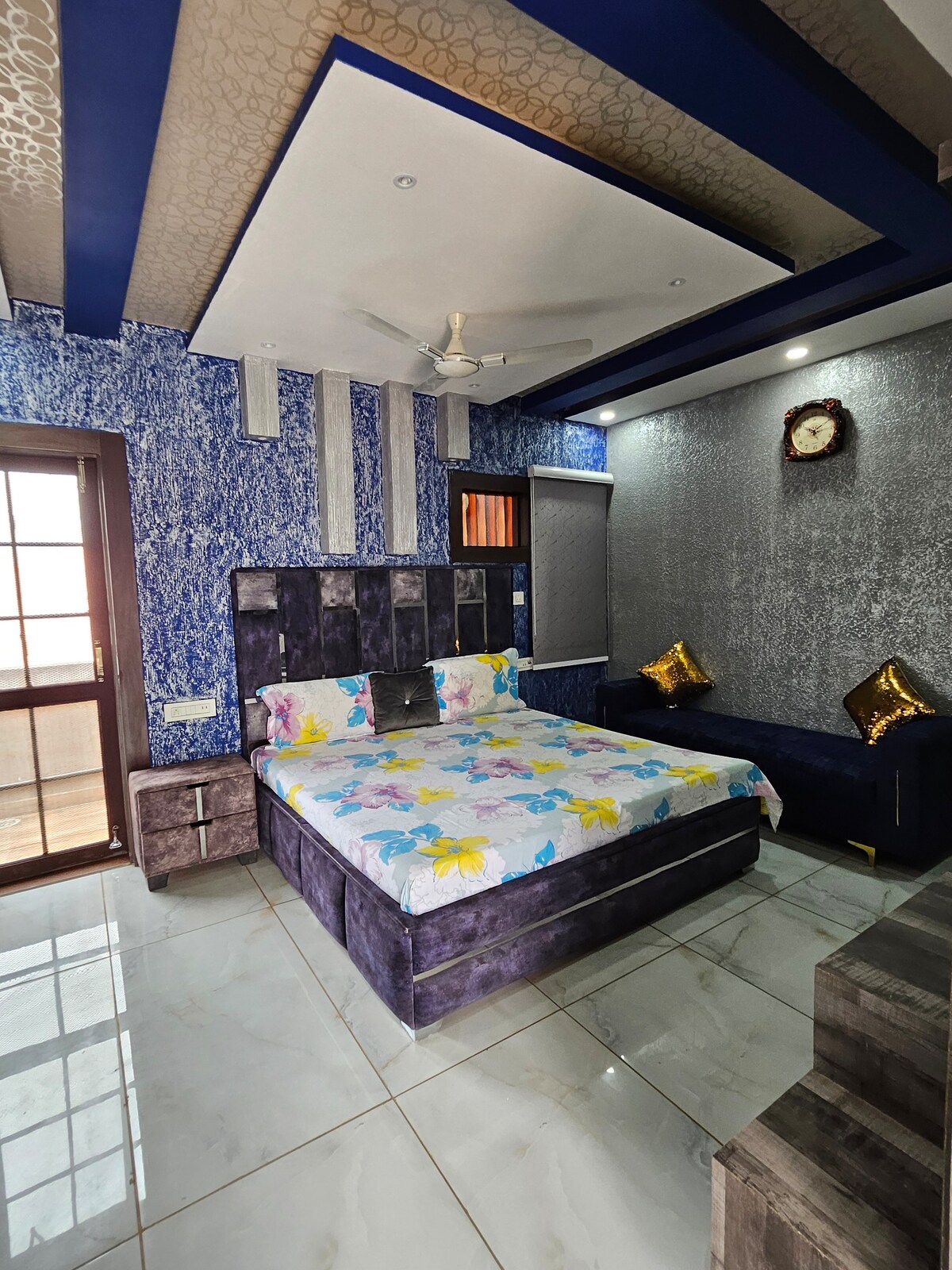 A bedroom is presented with a spacious bed adorned in a floral-patterned coverlet. Walls feature a mix of blue hues and metallic accents. Natural light filters through a window, complemented by a stylish clock on the wall. A cozy seating area includes a dark sofa with decorative cushions.