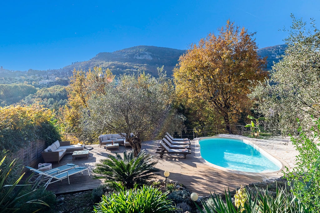 A serene outdoor space features a shaped pool surrounded by sun loungers and cozy seating areas. Lush Mediterranean plants and olive trees provide a natural backdrop, while the mountains in the distance enhance the tranquil atmosphere. The scene is complemented by autumn foliage.