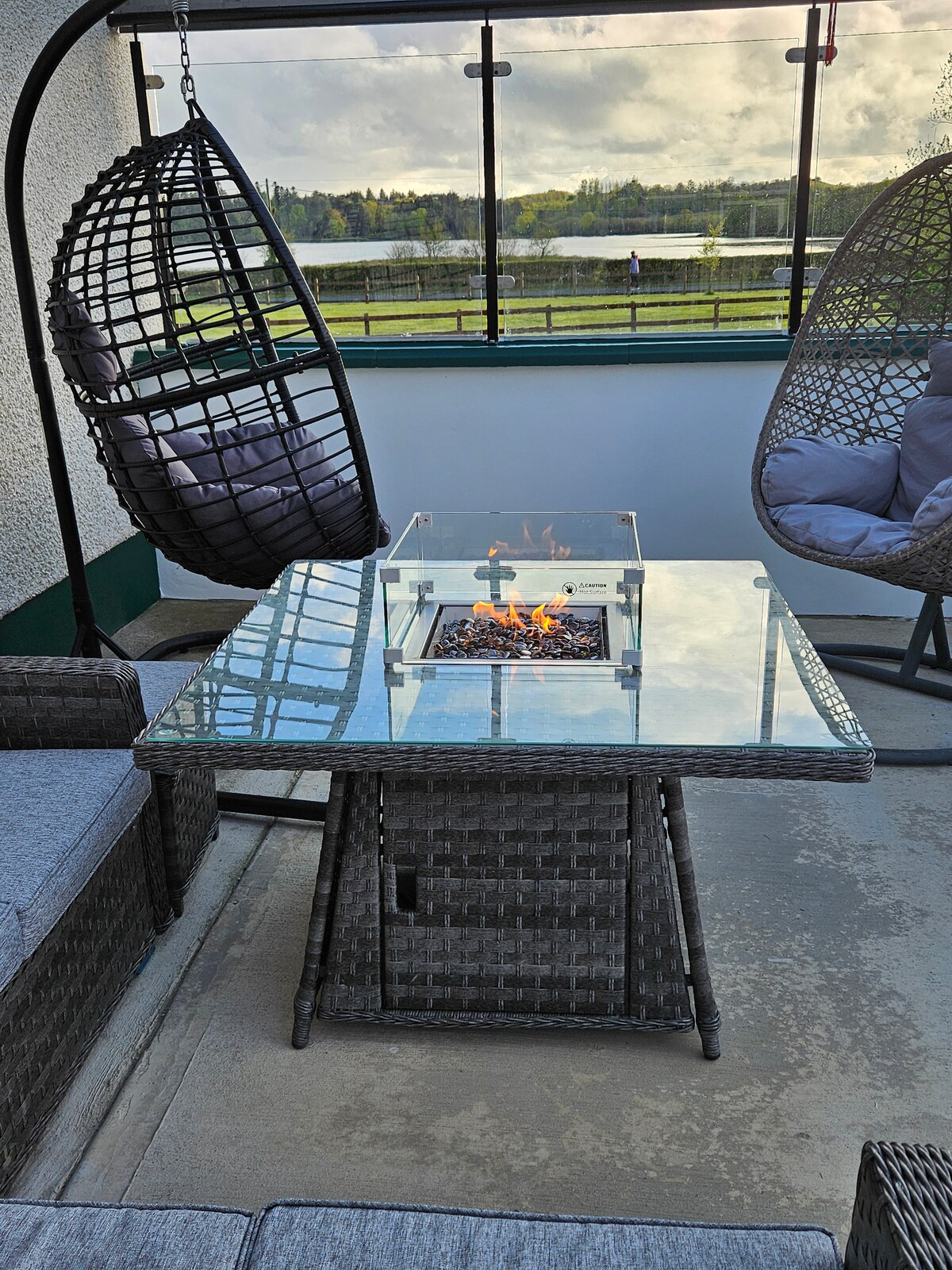 A cozy outdoor seating area features two hanging chairs and a modern table with a glass top, centrally located above a fire feature. A view of the landscape beyond is visible, with the serene water reflecting the light.