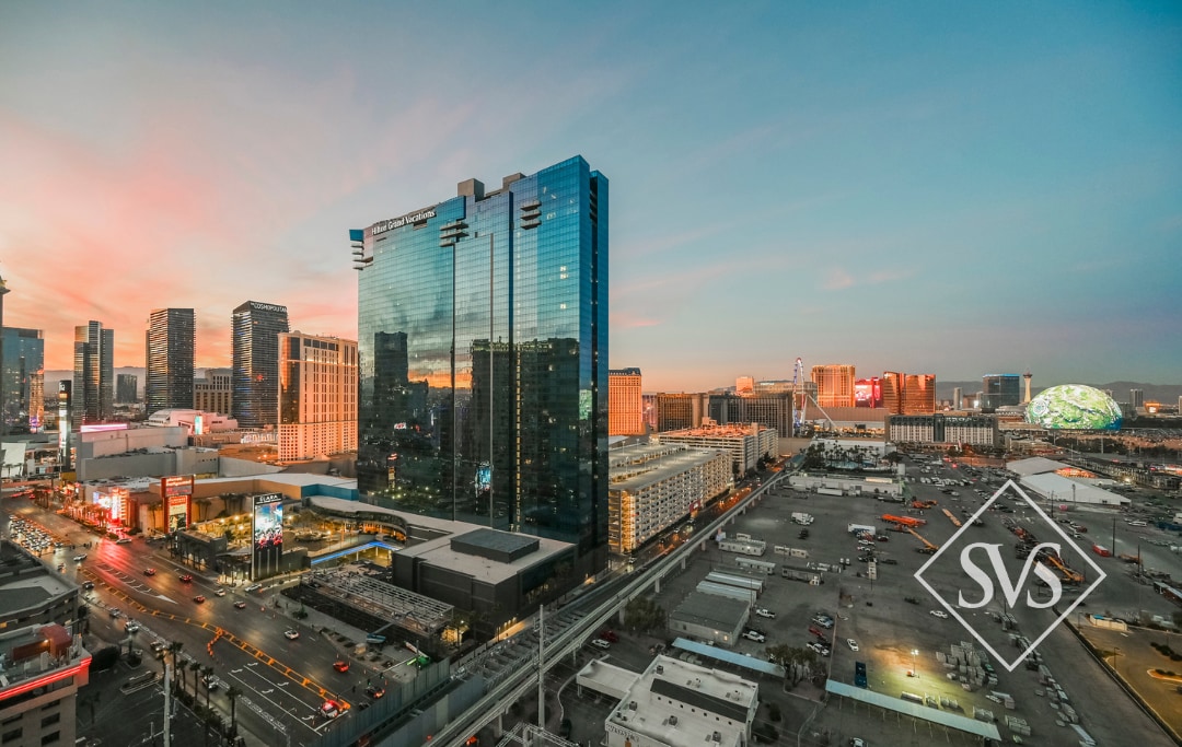 An expansive view showcases the Las Vegas Strip at dusk, with vibrant sky colors reflecting off modern high-rise buildings. Streets are bustling with traffic, while illuminated signs highlight the lively atmosphere of the city. The surrounding areas reveal expansive parking lots and additional developments.