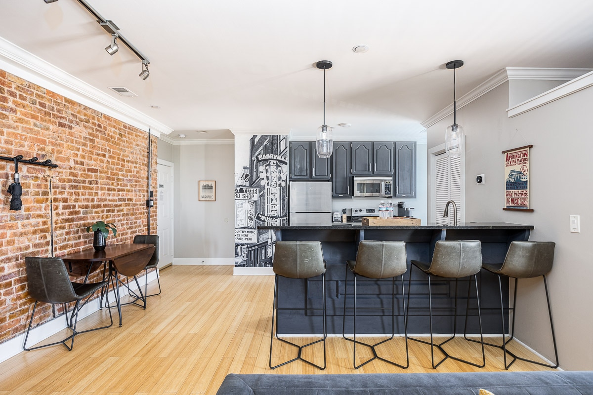 An open-concept living space features a brick accent wall and a modern kitchen with stainless steel appliances. A stylish black bar with four high stools is situated in front of the kitchen. A dining area with a wooden table is seen in the foreground.