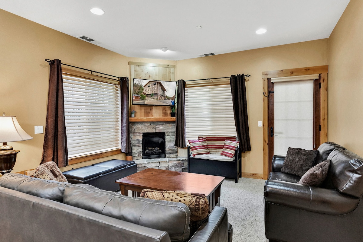 A comfortable living area features a stone fireplace as a focal point. Two large windows with layered treatments allow natural light, complemented by soft seating arrangements. A wooden coffee table sits centrally, and decorative elements add a homey touch.