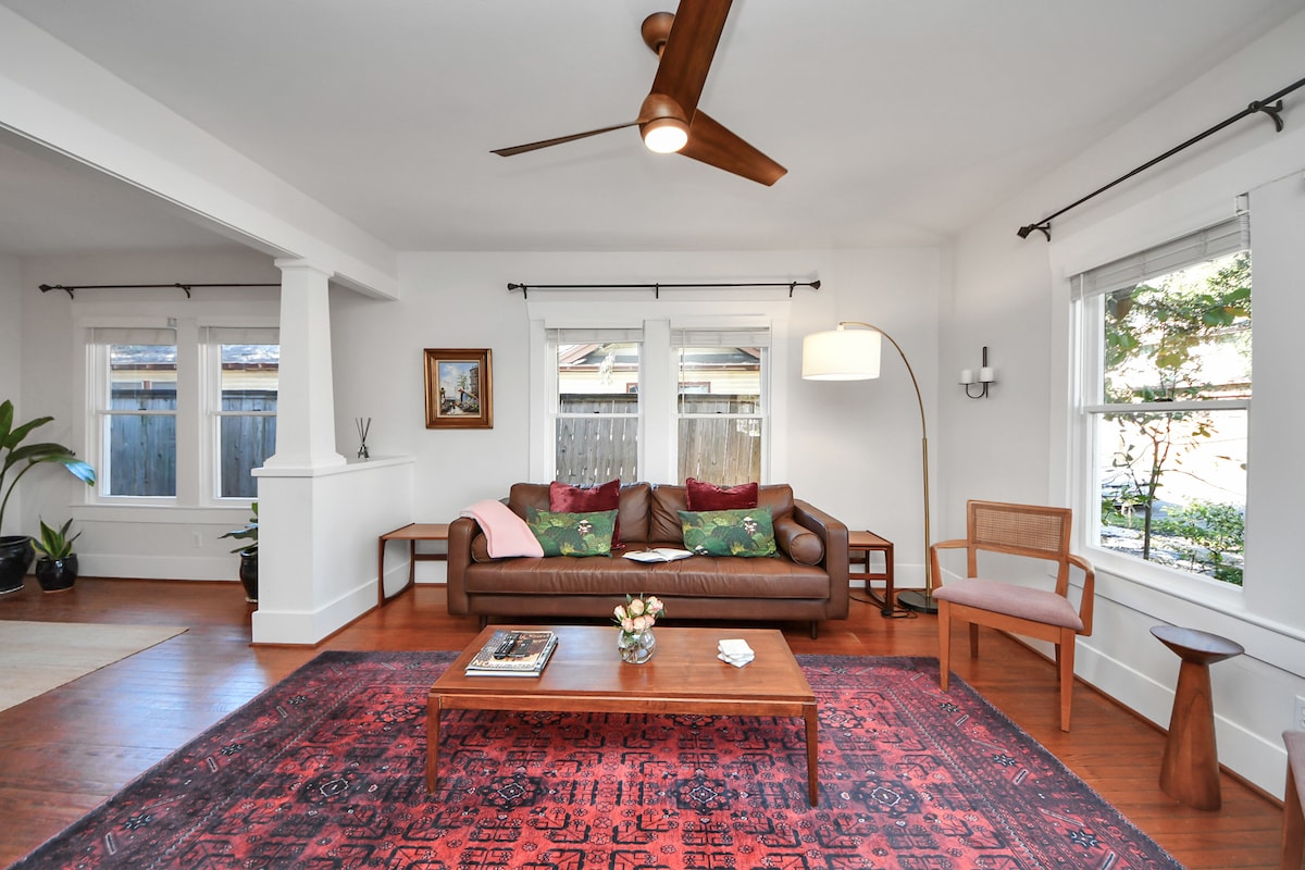 A comfortable living area features a leather sofa adorned with colorful cushions, situated on a patterned rug. Natural light enters through two windows, framed by soft curtains, and a ceiling fan is positioned overhead. A small coffee table displays books and a floral arrangement.