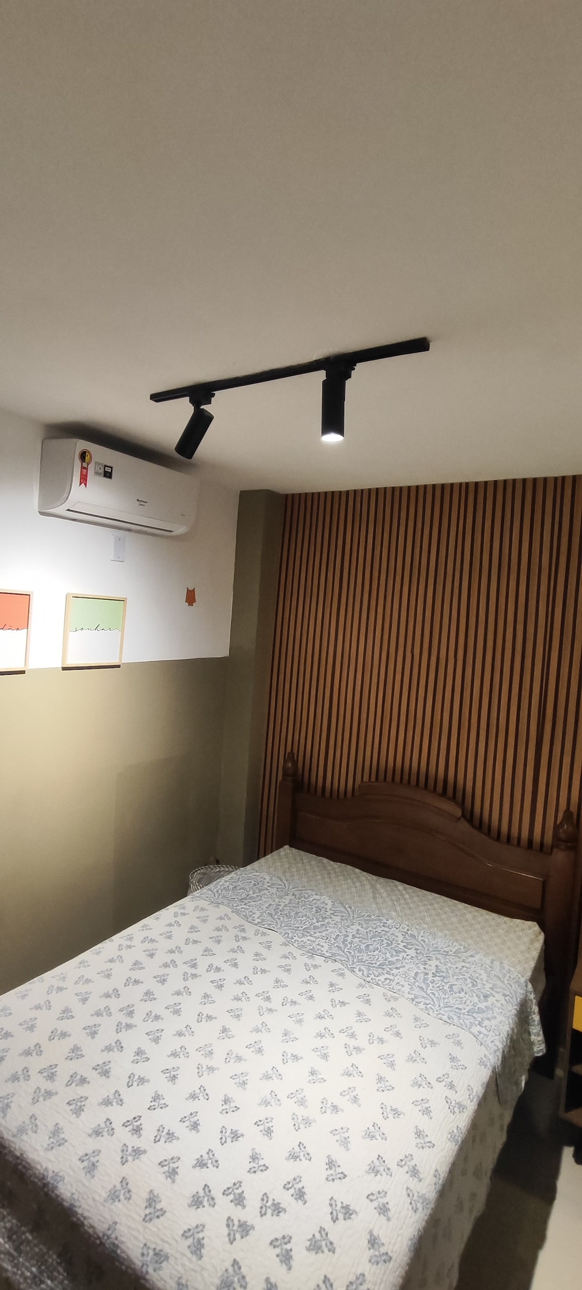 A compact bedroom features a wooden bed with a decorative blanket, complemented by textured wall panels. Above, track lighting provides illumination, while an air conditioning unit is mounted on the wall, ensuring comfort. Colorful wall art adds a touch of personality.