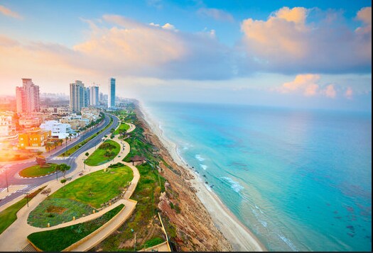 A panoramic view reveals a coastal landscape, showcasing a sandy shoreline meeting calm blue waters. The skyline is marked by modern high-rise buildings, while a winding path outlines the waterfront, surrounded by green spaces and walking areas, all under a soft, pastel sky.