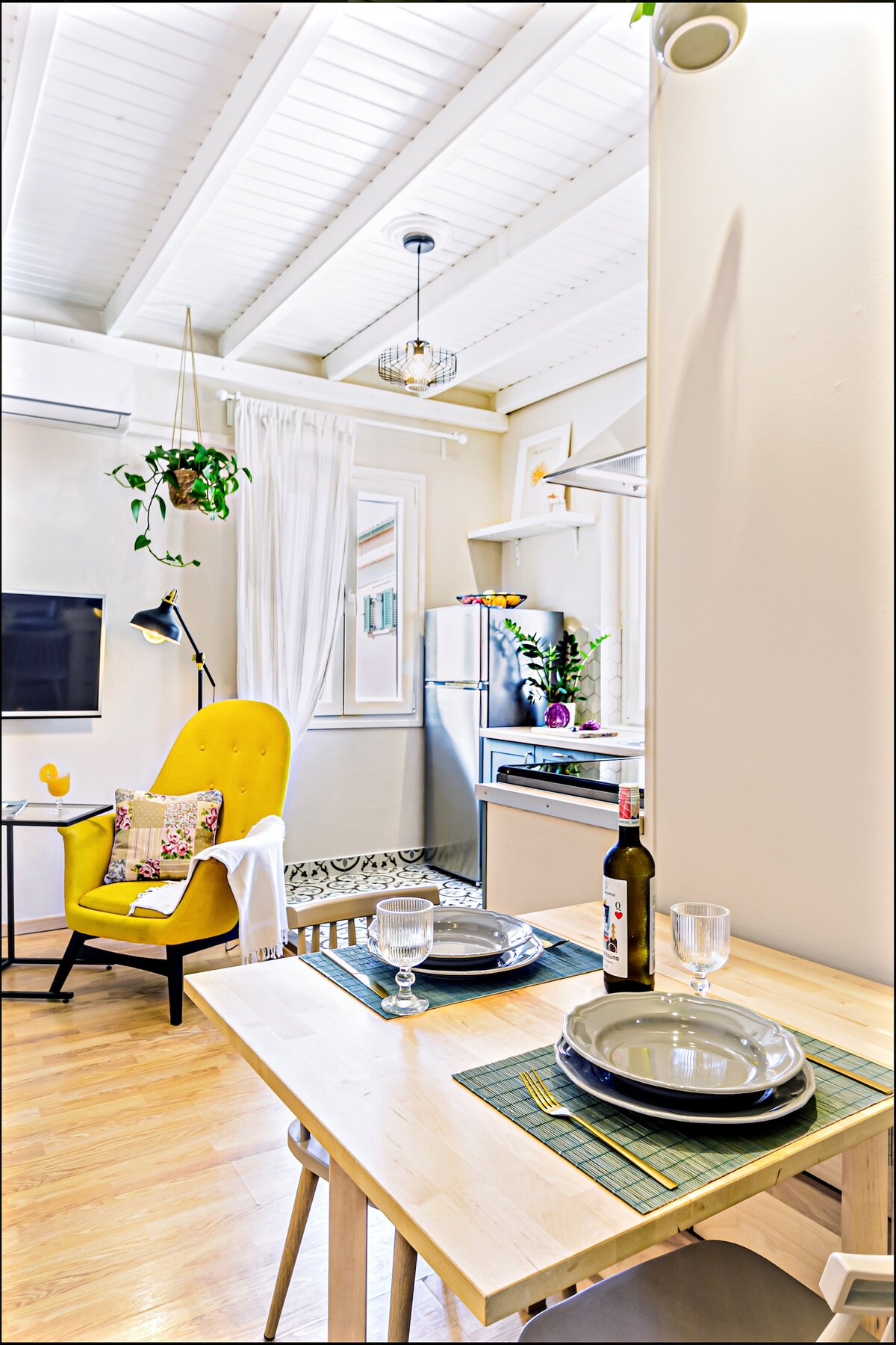 A bright dining area features a wooden table set with plates and glasses. A plush yellow chair adds a pop of color, while a well-lit kitchen area can be seen in the background. Natural light streams through the window adorned with sheer curtains.