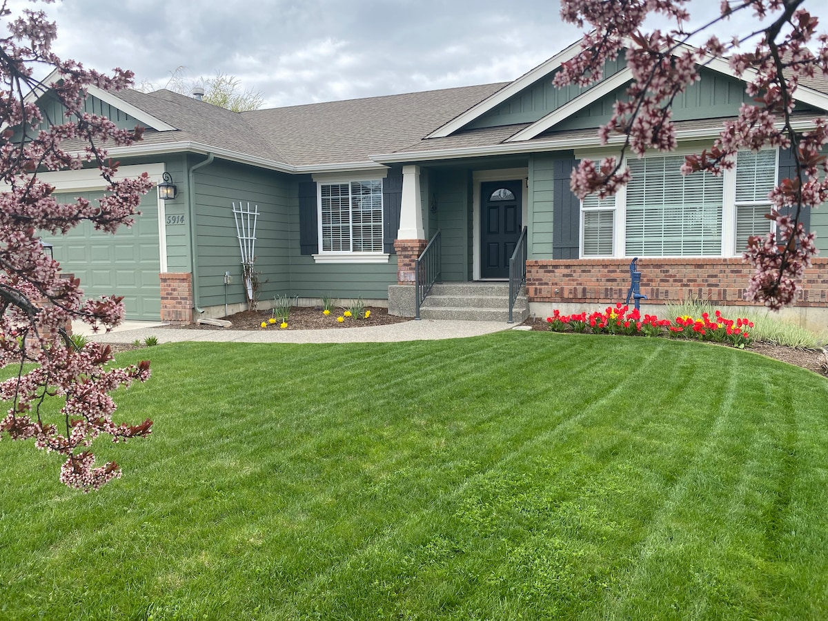 A well-maintained single-story home features a welcoming front porch with decorative plants, surrounded by lush green grass. Vibrant flower beds with yellow and red blooms add color to the landscape, while a gray driveway leads to the garage on the left.