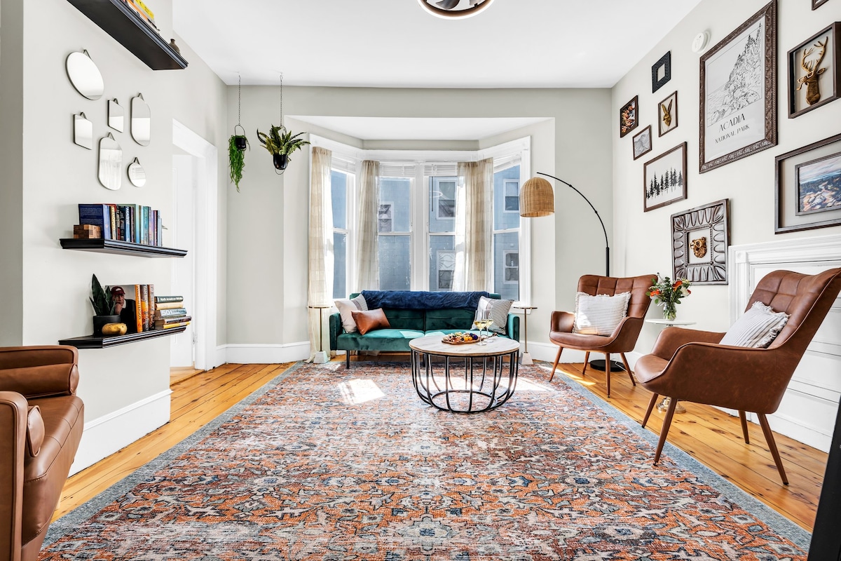 The open living area features a comfortable blue sofa and two brown accent chairs arranged around a round coffee table. Natural light fills the space through large bay windows, illuminating the area rugs and wall decor, creating a welcoming environment for relaxation.