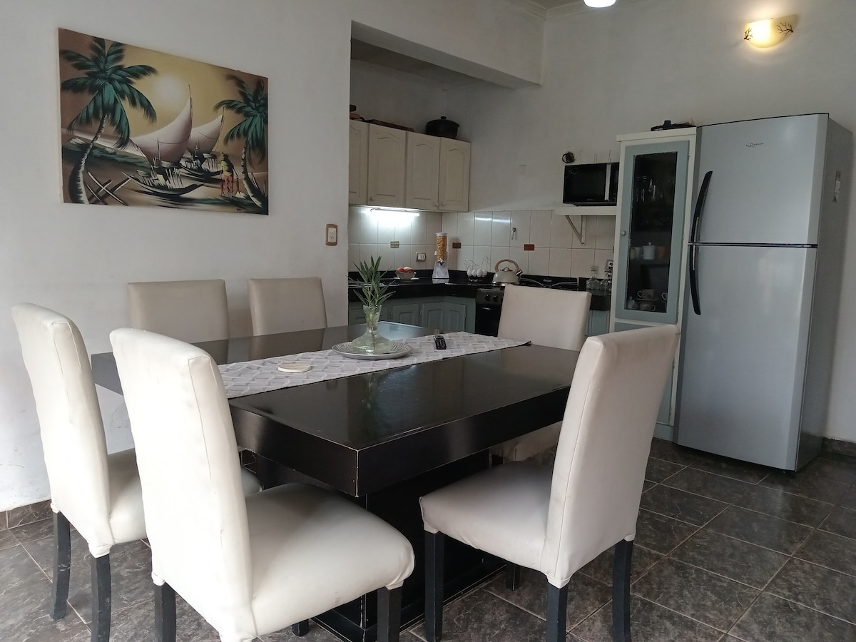 A bright dining area features a large black table surrounded by six light-colored upholstered chairs. A painting depicting a sailboat hangs on the wall, adding a touch of color to the space. A refrigerator and kitchen appliances are visible in the open kitchen area.