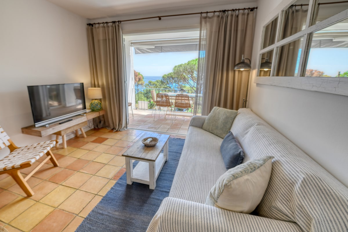 A lounge area features a comfortable couch with decorative pillows, alongside a wooden coffee table. A television is positioned on a stand. Large glass doors open to a terrace, revealing views of greenery and the sea beyond. Warm lighting illuminates the space.