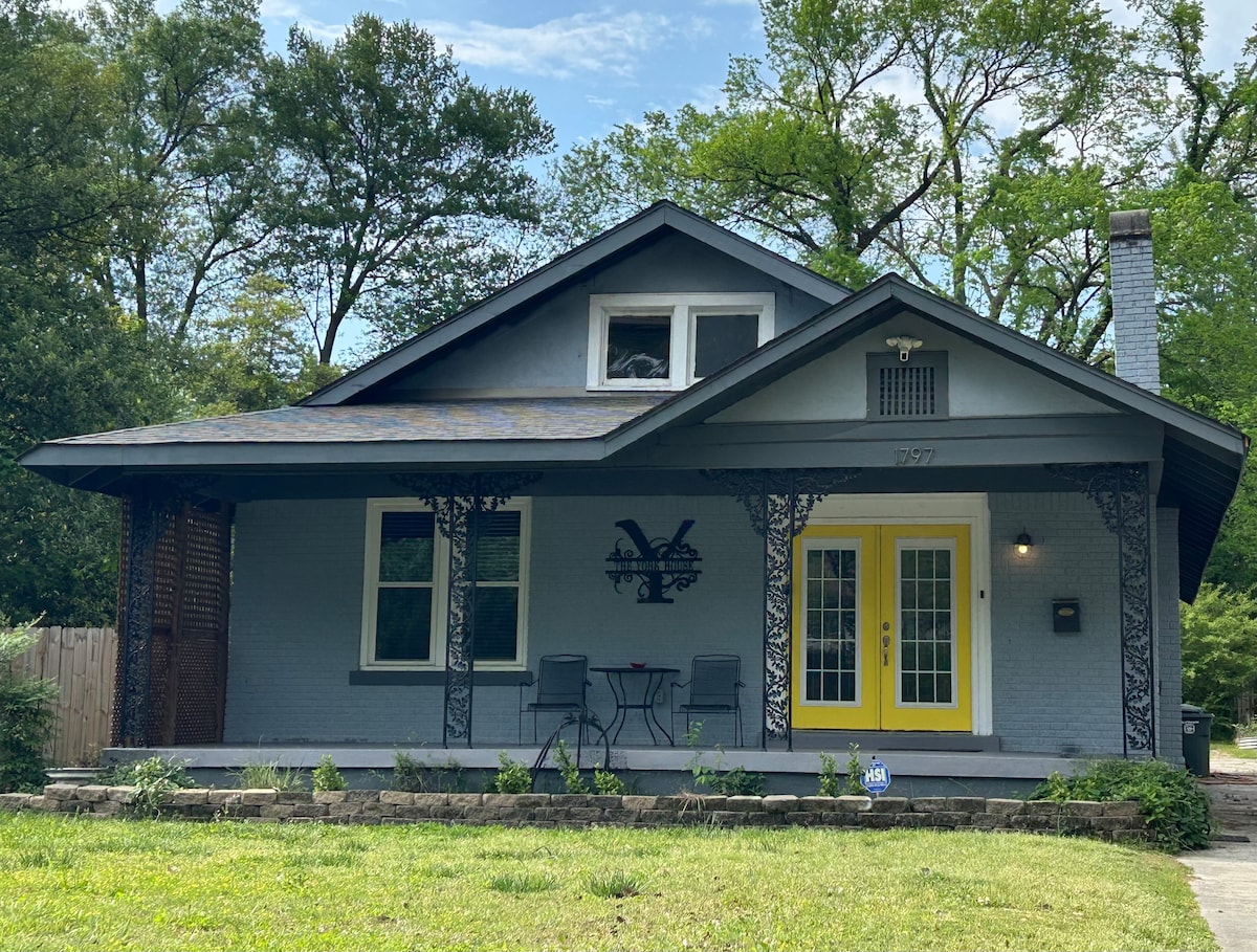 A charming house with a grey exterior displays a welcoming yellow front door. Two black chairs are positioned on the porch, surrounded by greenery. The porch is supported by intricate wooden columns, and a stone pathway leads to the entrance.