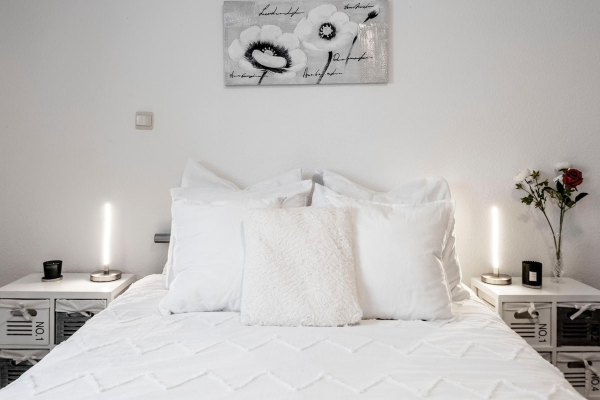 A well-lit sleeping area features a spacious bed adorned with multiple white pillows. On either side, minimalist bedside tables hold decorative candles and slender lamps. A flower arrangement adds a touch of nature, complemented by a simple floral artwork above the bed.