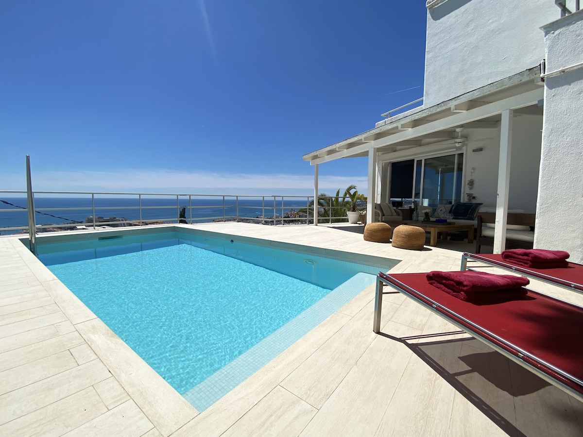 An inviting outdoor space features a clear swimming pool surrounded by a sun terrace. Loungers are positioned nearby, with plush towels neatly arranged on them. The ocean view stretches into the distance, complemented by abundant natural light from the clear sky.