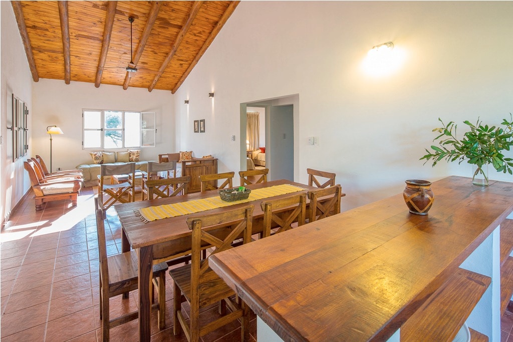 A spacious dining area is highlighted by a wooden table surrounded by multiple chairs. Natural light enters through a window, illuminating the warm wooden ceiling and creating a welcoming ambiance. A small decorative bowl is placed on the table, adding a touch of detail.