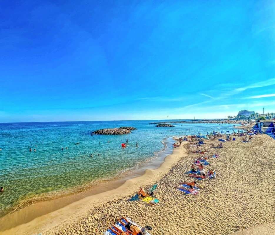A sandy beach is depicted under a clear blue sky, with gentle waves lapping at the shore. Sunbathers are seen lounging on towels near the water's edge, while people enjoy leisurely activities in the sea. Rocky formations are visible in the distance.