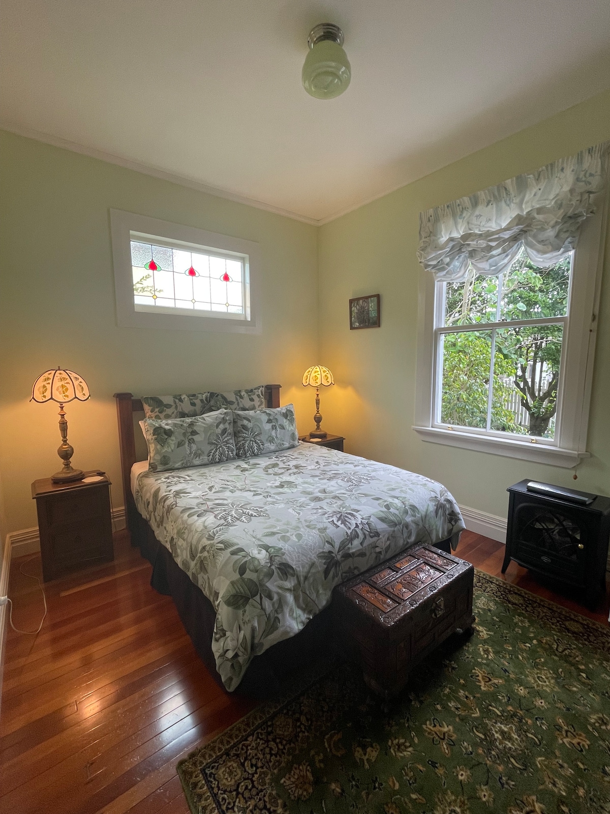 A cozy bedroom features a spacious bed adorned with floral bedding and two bedside lamps. Natural light filters through a window with decorative stained glass, while a vintage trunk sits at the foot of the bed. A patterned rug adds warmth to the hardwood floor.
