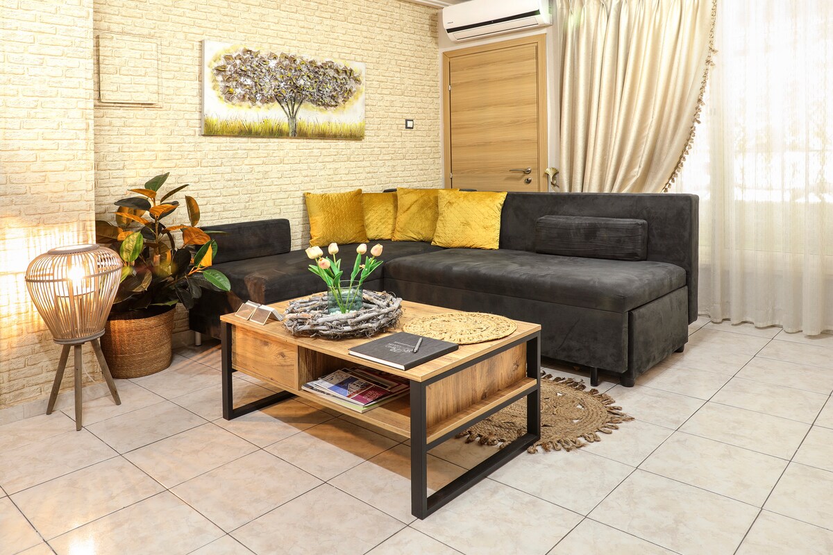 A cozy living area features a dark sectional sofa adorned with mustard yellow pillows. A wooden coffee table sits in front, displaying magazines and a decorative bowl. A decorative plant, woven rug, and light fixtures add warmth to the space, enhanced by natural light from nearby windows.