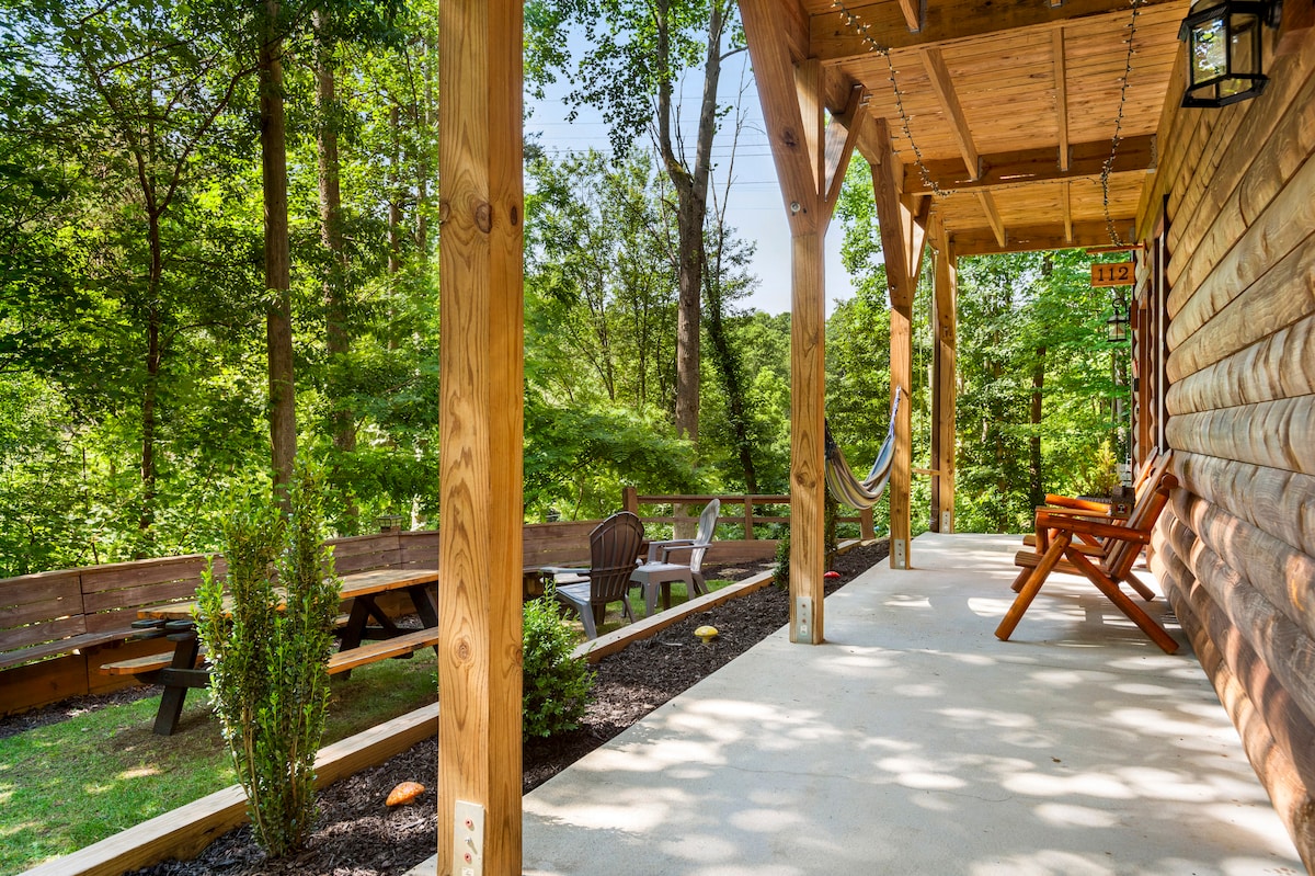 A covered porch is framed by wooden beams, providing a view of the surrounding greenery. Comfortable seating options include rustic rocking chairs and a hammock. A picnic table and additional seating are visible in the outdoor area, with landscaped plants alongside a concrete walkway.