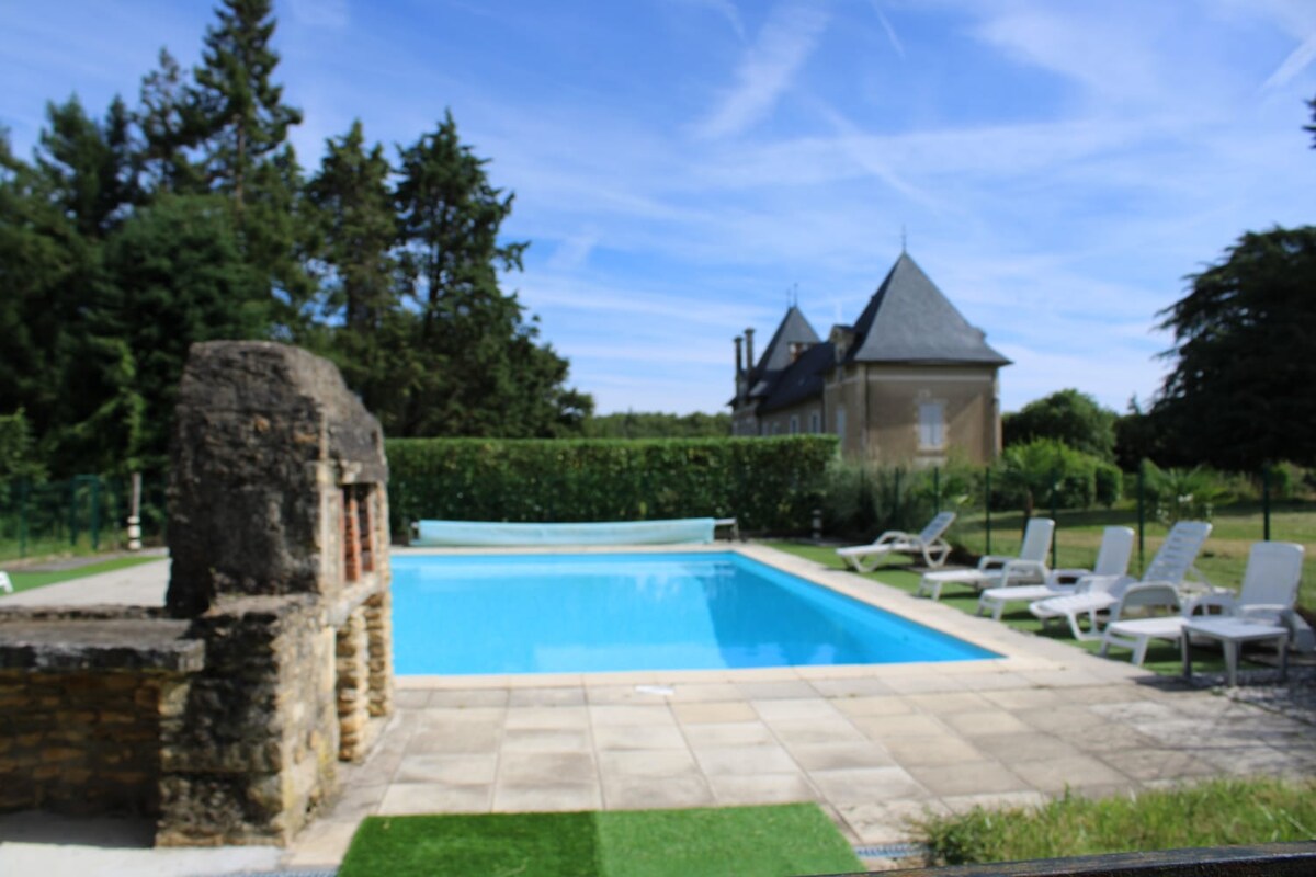 An outdoor pool is located within a spacious deck area, surrounded by lounge chairs for relaxation. A stone fireplace is visible alongside the pool, adding character. Lush greenery and trees frame the pool area, providing a natural backdrop.