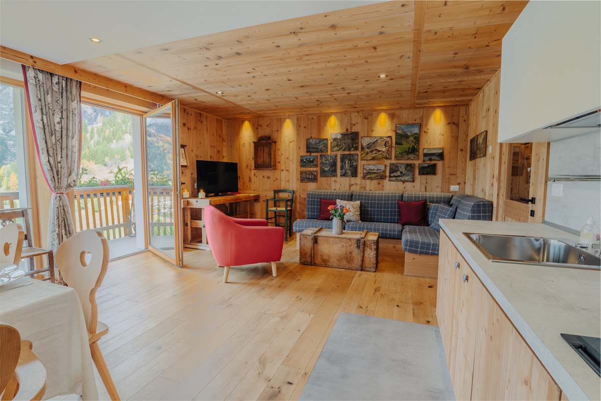 A spacious living area features wooden walls and a cozy seating arrangement with a vibrant red chair and a blue couch. Natural light floods the space through large windows, and several framed artworks adorn the walls. A dining table is positioned nearby, enhancing the inviting atmosphere.