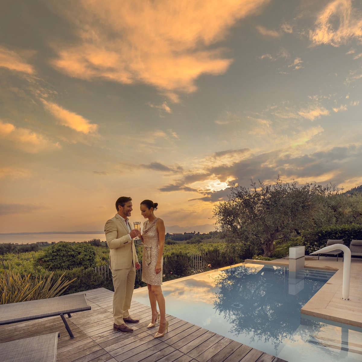 A serene pool area is featured, reflecting the colorful sunset and surrounding greenery. Two guests are seen enjoying drinks, while a gently sloped lawn leads toward olive trees. The modern wood deck offers space for relaxation, complementing the tranquil atmosphere.