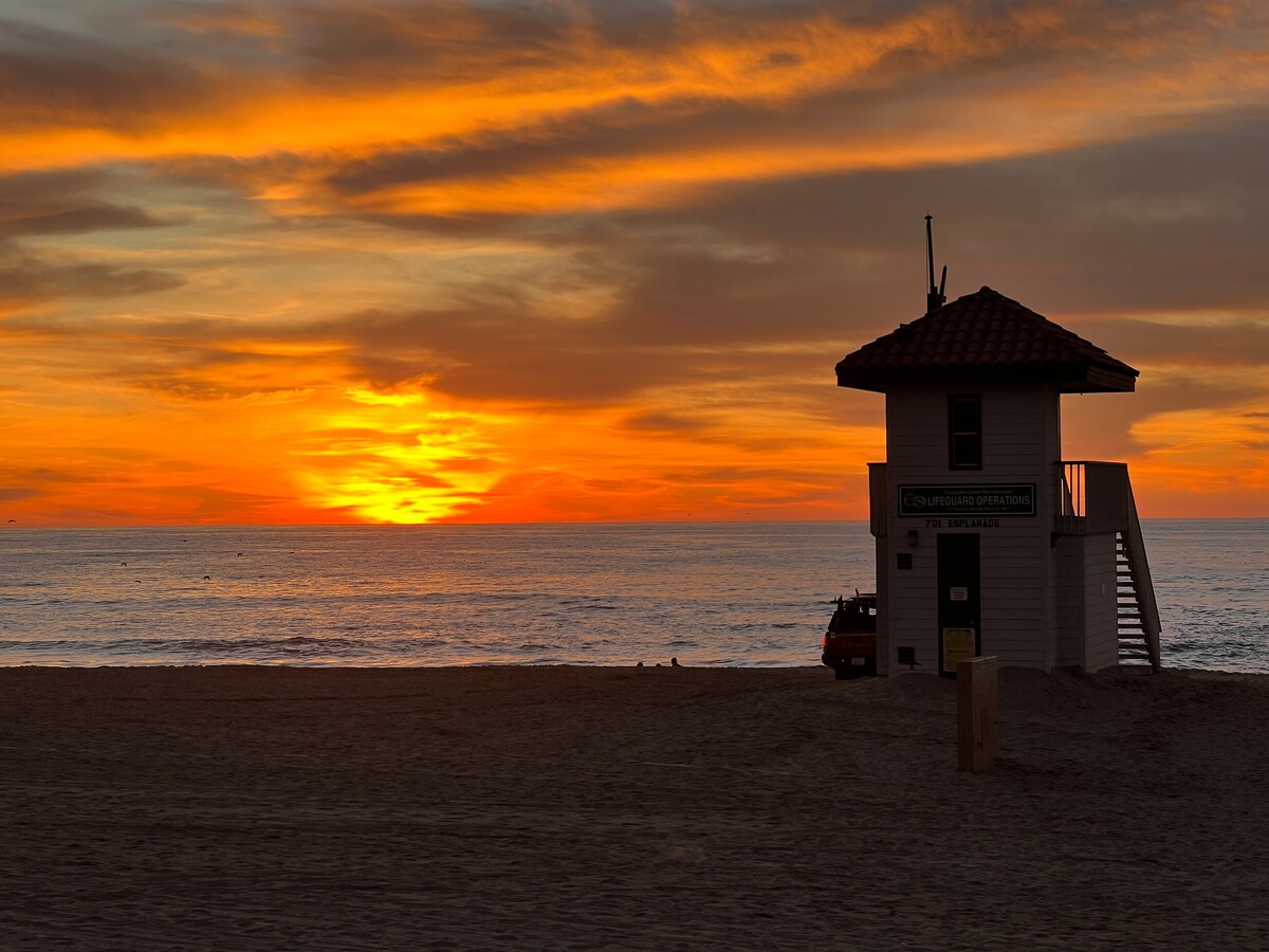 A lifeguard station stands along the sandy beach, silhouetted against a vivid sunset. Warm hues of orange and yellow blend into the sky, reflecting on the calm ocean waters. The horizon showcases a serene and inviting coastal landscape, enhancing the tranquil evening ambiance.