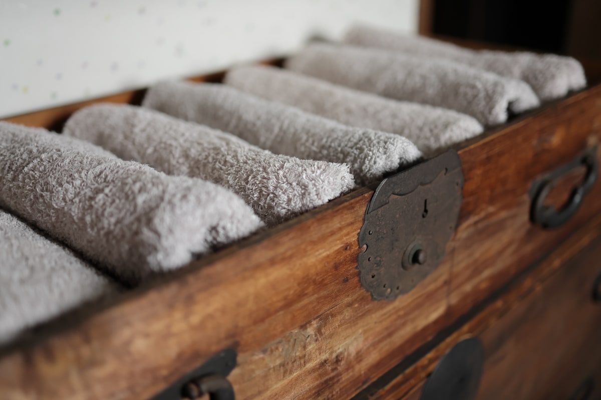 A wooden chest holds neatly rolled gray towels, creating an organized and inviting presentation. The texture of the towels is easily discernible, providing a sense of comfort and relaxation for guests. The interior features subtle patterns in the background.