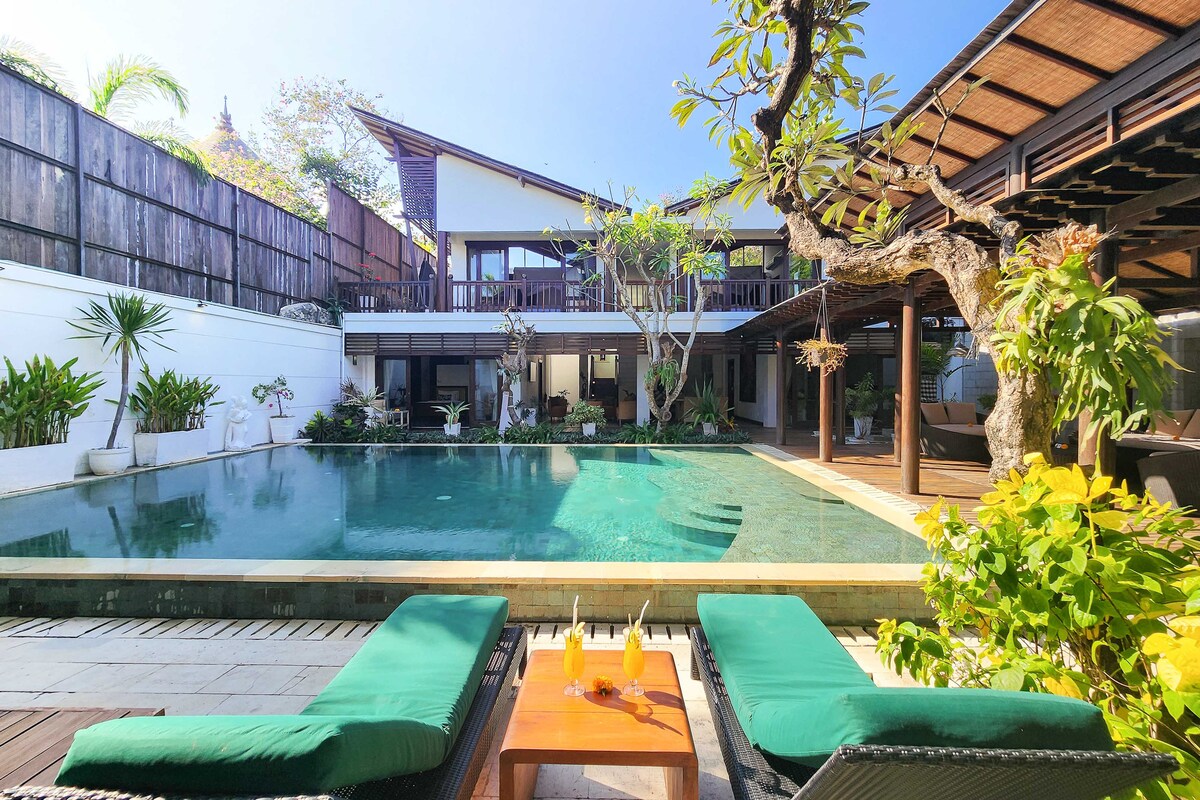 A spacious pool area is depicted, featuring a large, inviting pool surrounded by lush greenery. Two lounge chairs with green cushions are positioned on the deck, accompanied by a wooden table holding refreshments. The villa's architecture, with a blend of traditional and modern elements, is visible in the background.