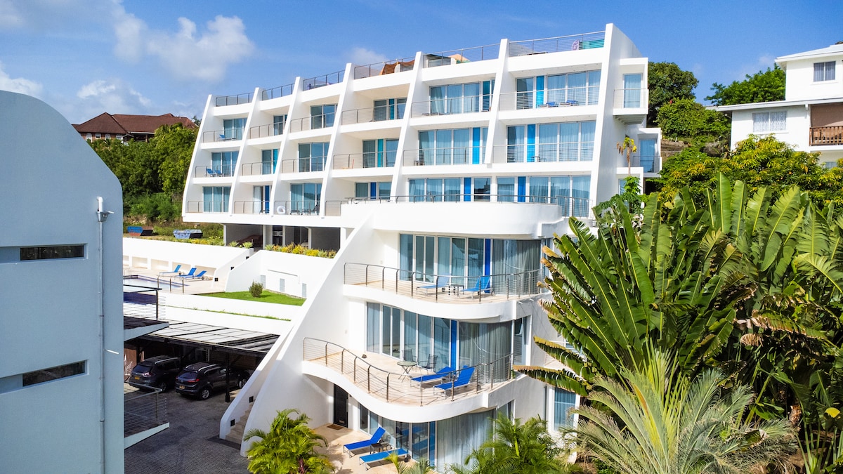 A contemporary condominium complex is showcased, featuring modern architecture with multiple levels. Expansive glass windows dominate the facade, allowing natural light to fill the spaces. Sun loungers are positioned on the balconies, while lush greenery surrounds the property, enhancing the inviting outdoor environment.