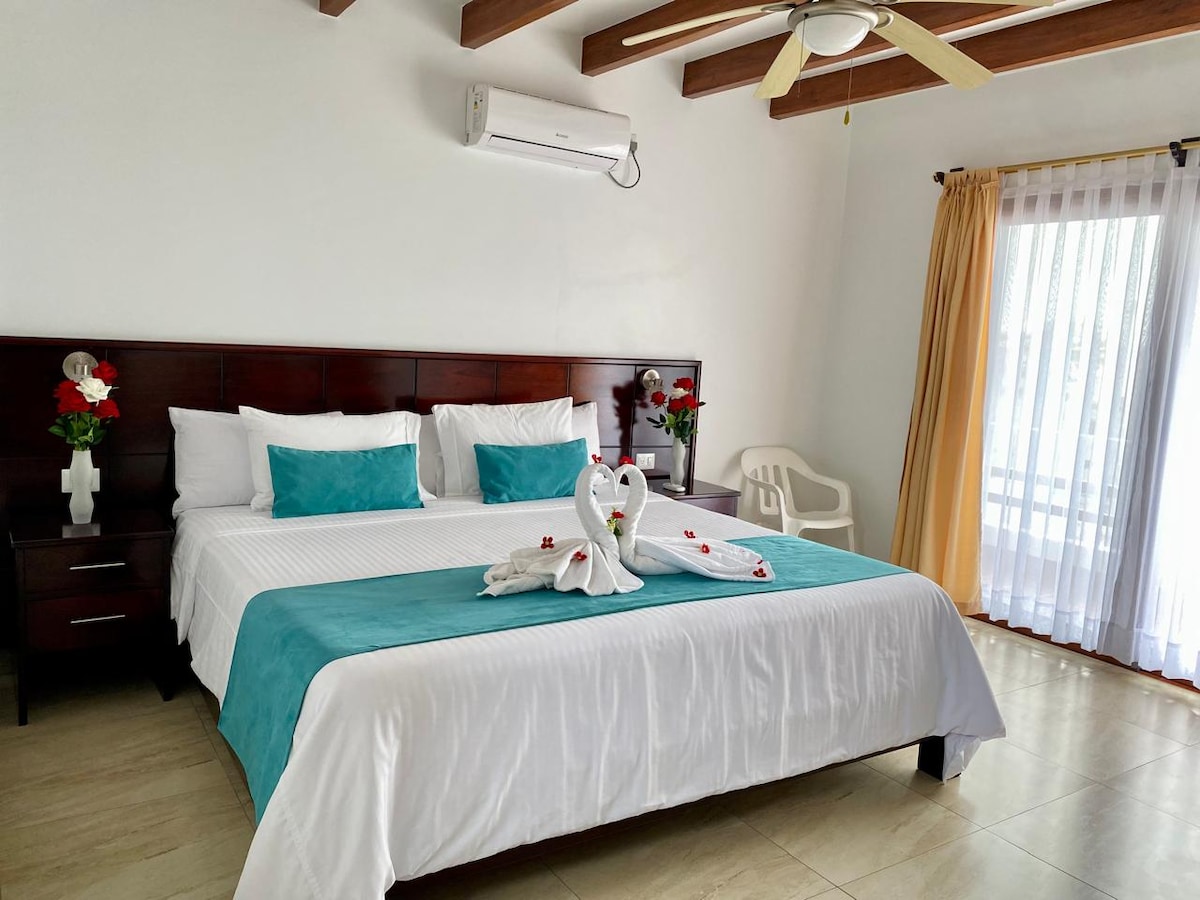 A spacious bedroom features a large bed dressed in white linens with a turquoise accent blanket. Soft light enters through sheer curtains, highlighting the wooden ceiling beams. Nightstands with floral arrangements complement the calming environment, and a ceiling fan provides added comfort.