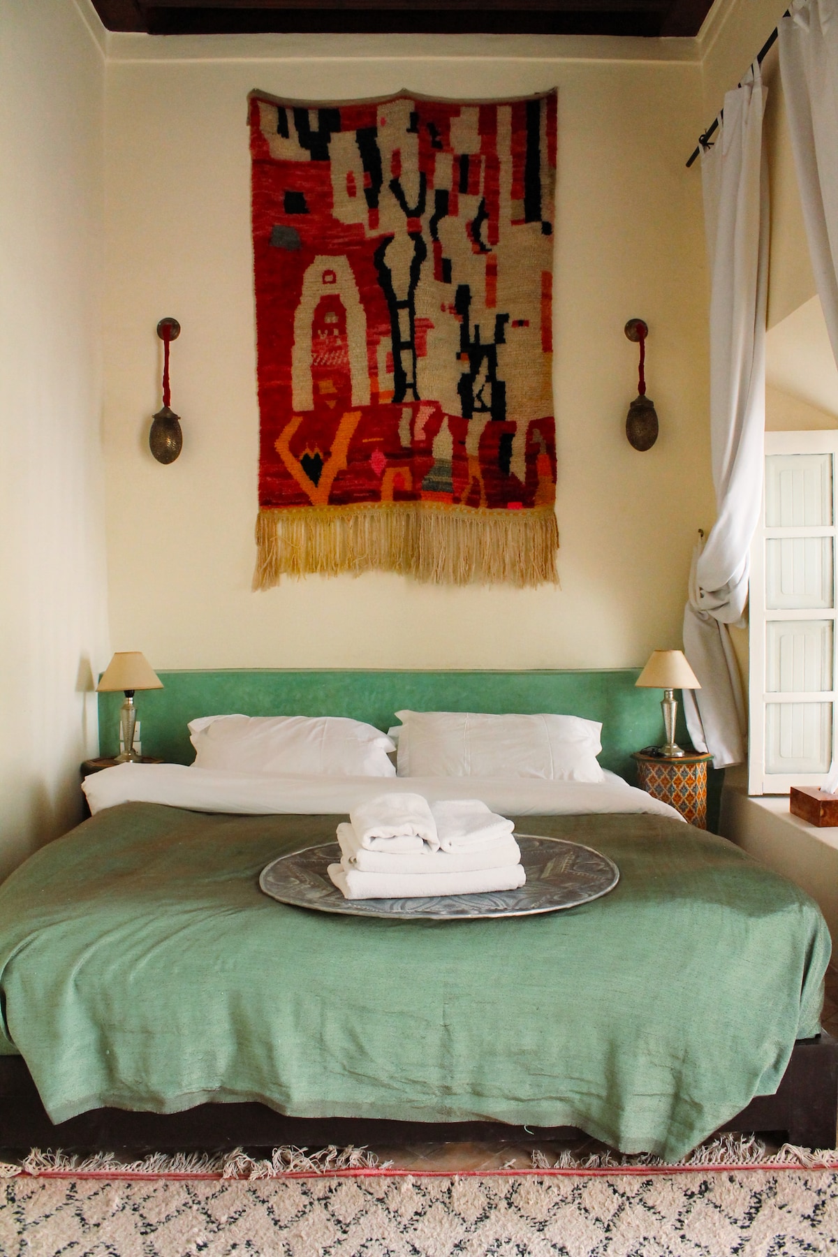 A calming bedroom is featured, with a spacious bed dressed in soft white linens and a muted green throw. A decorative wall hanging adds texture above the headboard. Two bedside lamps provide gentle light, and natural light enters through an open window draped with sheer white curtains.