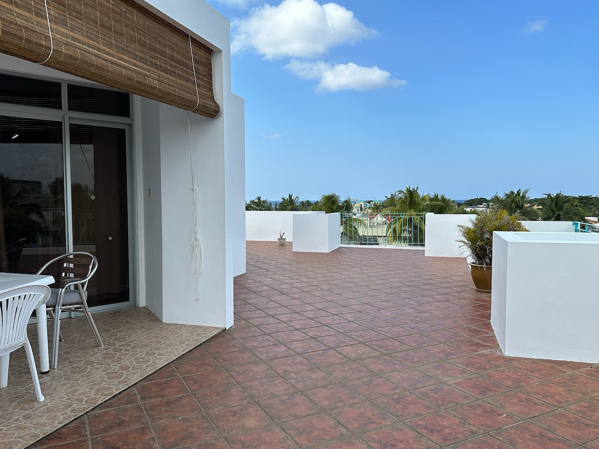 A spacious terrace area is presented, featuring a tiled floor and a dining set with white chairs. Panoramic views of the surrounding area are visible, including glimpses of greenery and the ocean beyond. The sky is partly cloudy, enhancing the outdoor ambiance.