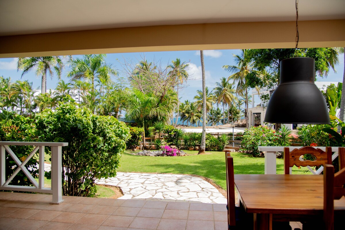 An outdoor terrace is visible, featuring a wooden dining table and chairs. Lush greenery surrounds the space, including tropical plants and vibrant flowers. A glimpse of the ocean can be seen in the background, framed by palm trees and a clear blue sky.