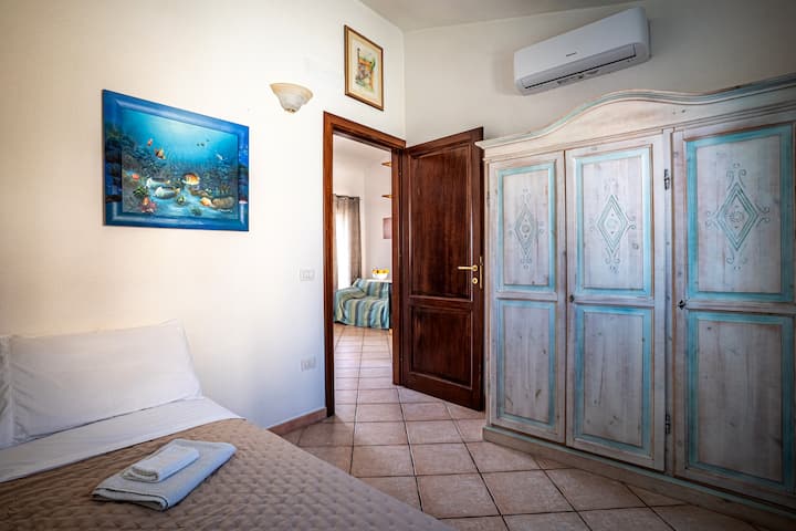 غرفة النوم التي تحتوي على سرير مفرد وخزانة وتكييف هواء، مع وجود غرفة المعيشة في الخلفية 