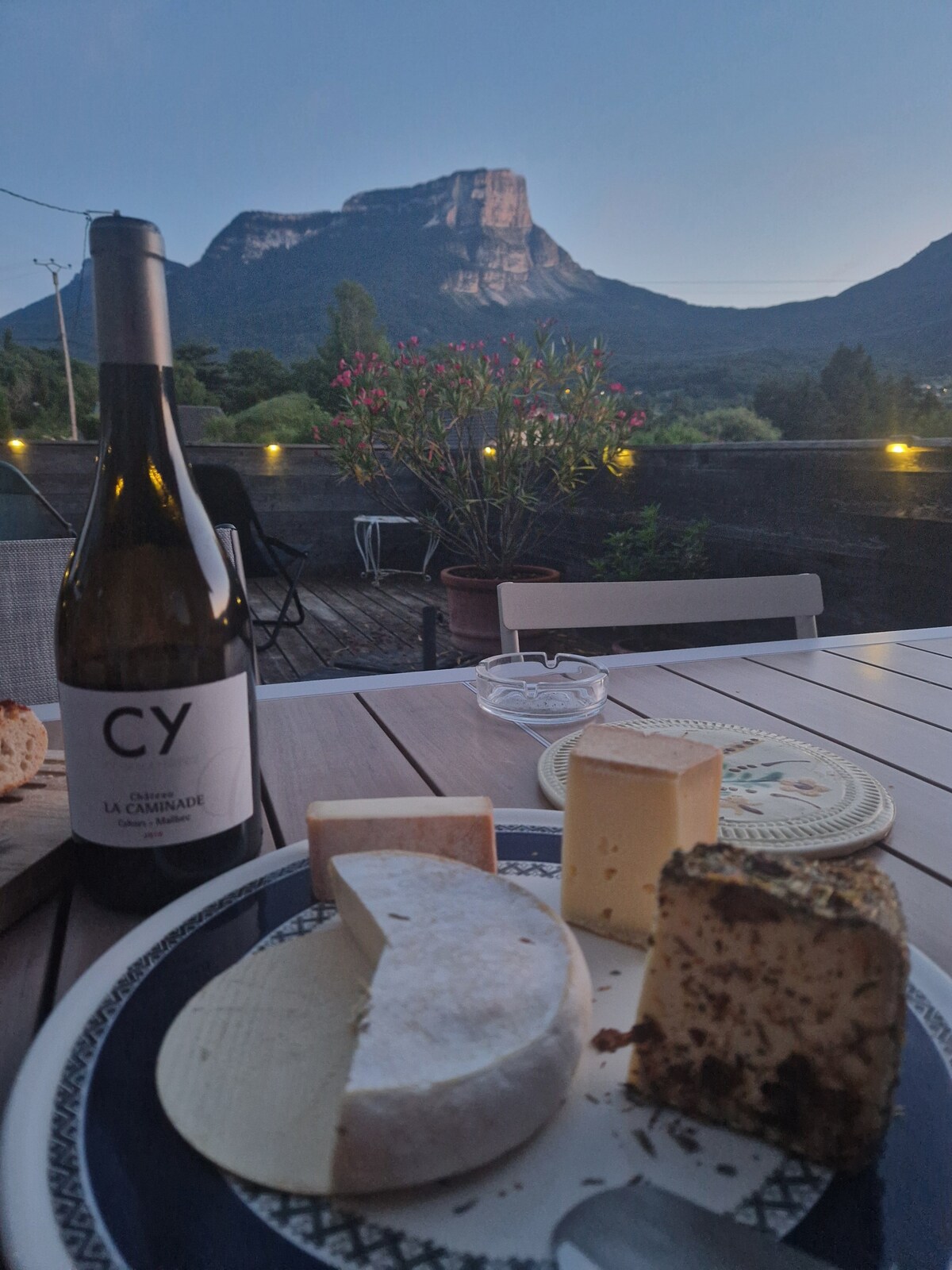 An outdoor dining table is set with a variety of cheeses and a bottle of wine, accompanied by a scenic mountain backdrop. Soft evening light highlights the landscape, with potted plants adding greenery to the space.