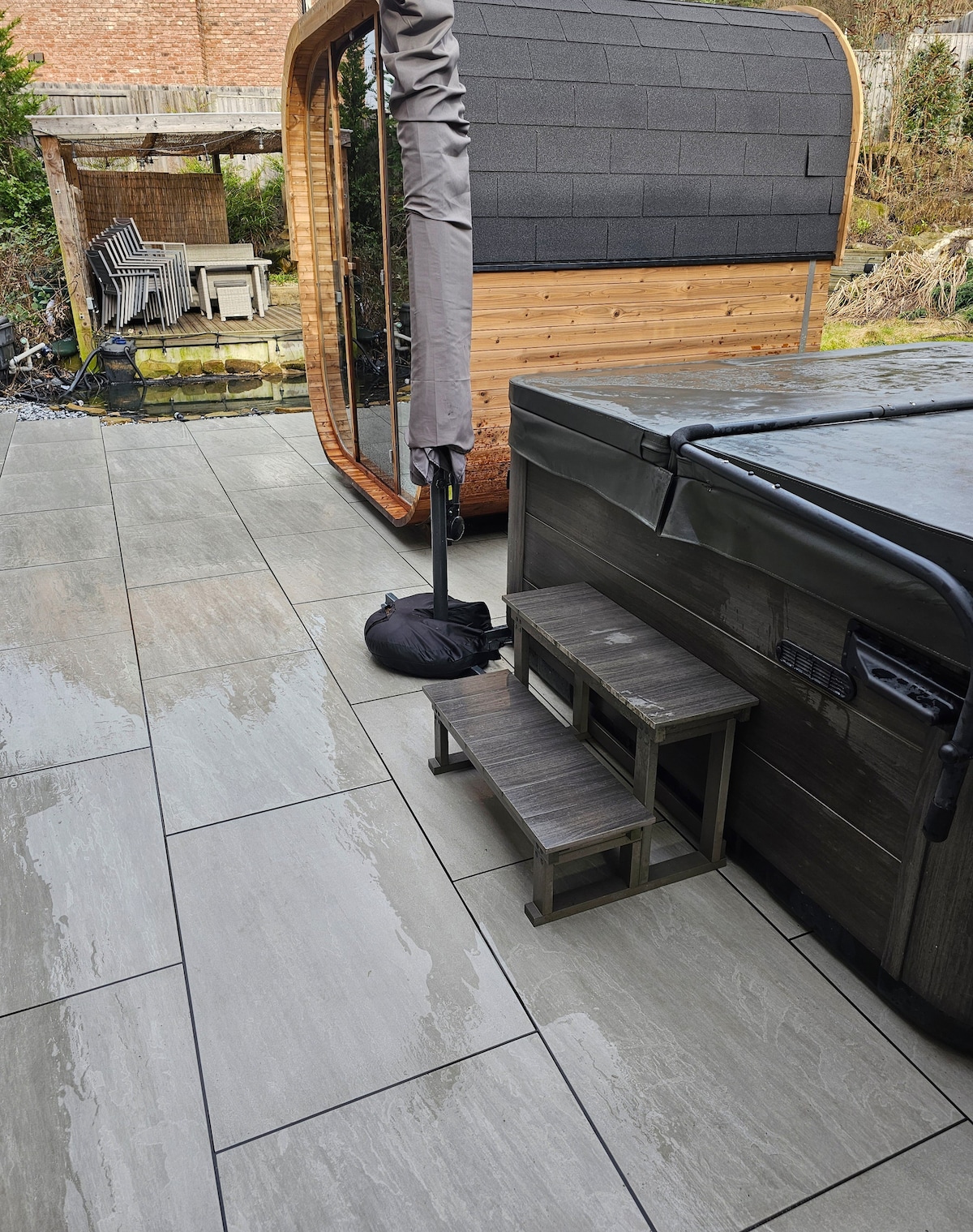 A spacious outdoor area features a hot tub alongside a wooden pod structure. Stone tiles lead to steps that access the hot tub, while a small table can be seen nearby. Natural greenery borders the patio, providing a serene backdrop.