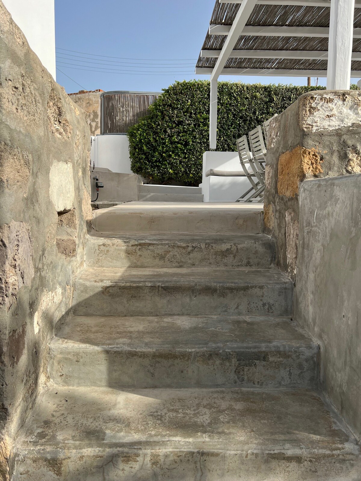 Stone steps lead upward, surrounded by rustic stone walls. A glimpse of greenery is visible at the top, where a shaded seating area awaits under a white canopy. Natural sunlight highlights the textured surfaces, creating a welcoming outdoor path.