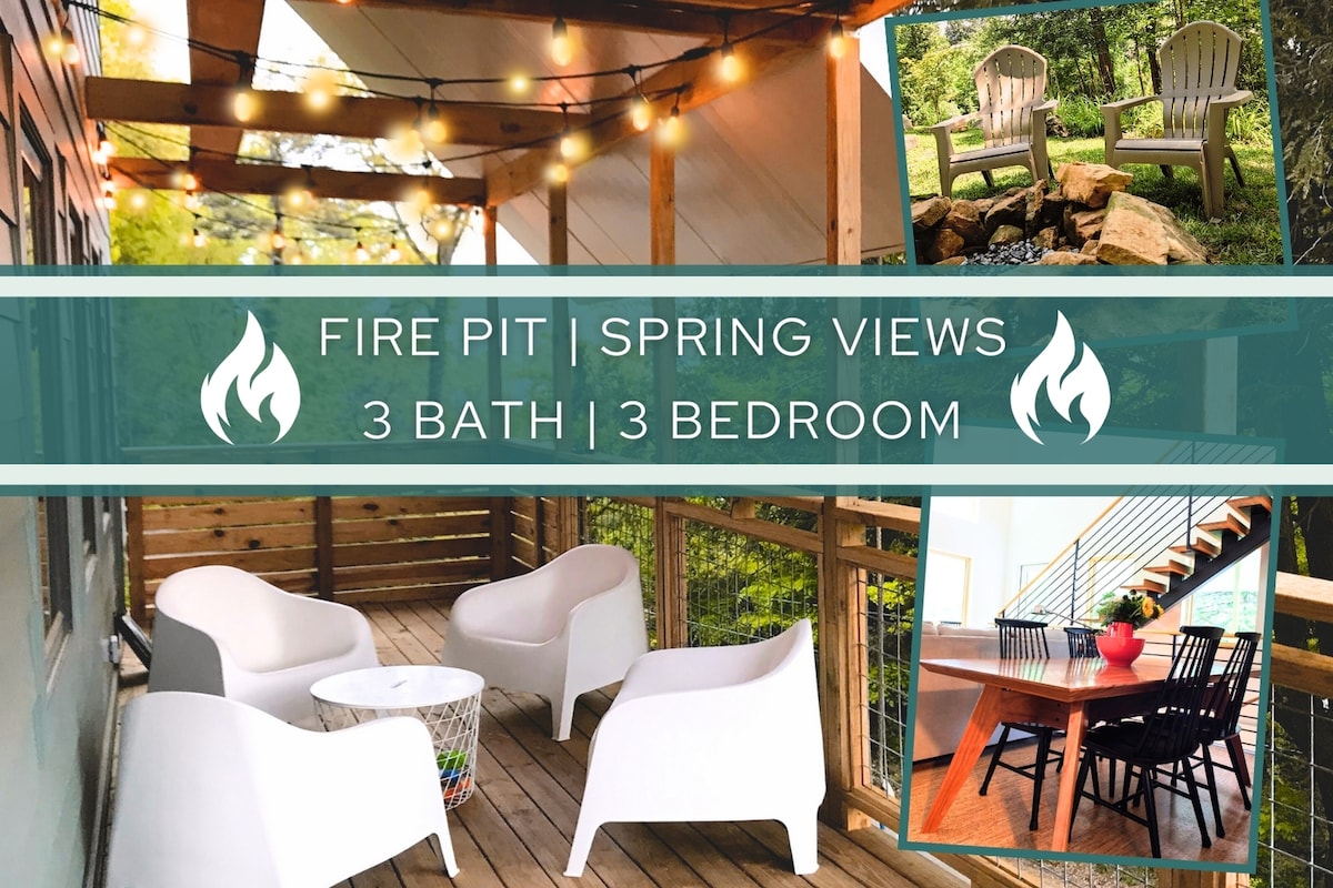 An outdoor seating area is showcased with white chairs under string lights. A fire pit is positioned nearby, surrounded by landscaping that includes rocks. The image also displays a dining table set for a meal, offering views of the green surroundings.