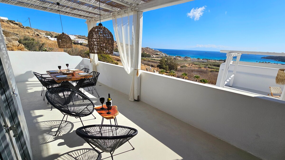 An expansive outdoor terrace features a dining table set for several guests, accompanied by comfortable chairs. Woven pendant lights hang above, enhancing the setting. A stunning view of the coastline and sea extends beyond, while clear blue skies complete the serene atmosphere.