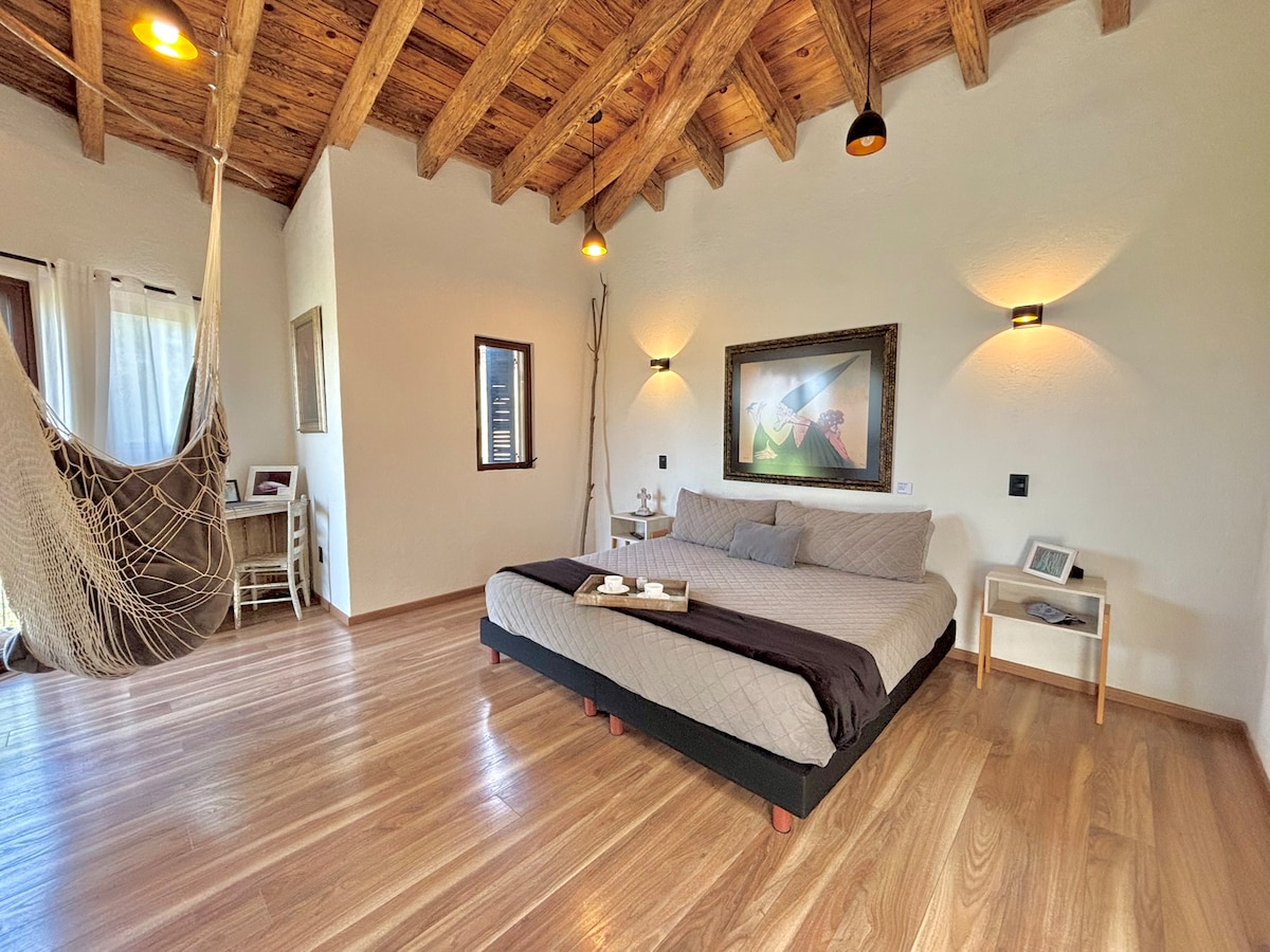 The spacious bedroom is adorned with wooden beams overhead and features a large bed with a neutral-colored quilt. A hammock is suspended in one corner, while natural light enters through large windows, illuminating the minimalist decor and wooden flooring.