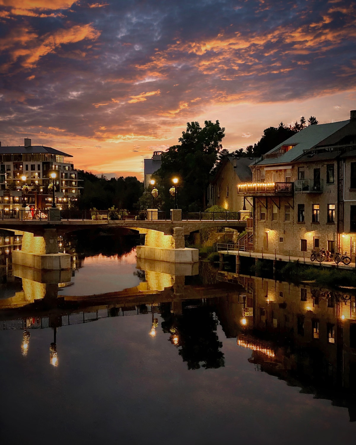 A serene river scene at sunset showcases reflections of historic buildings and a bridge adorned with street lamps. Warm light from nearby structures enhances the inviting atmosphere of the area, while streaks of orange and purple paint the evening sky.