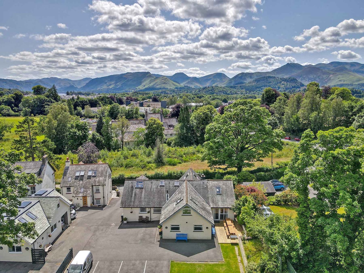 An aerial view captures the Borrowdale View House among lush greenery, with a spacious parking area visible. The surrounding landscape features rolling hills and a clear sky, creating a serene backdrop that reflects the natural beauty of the Lake District.