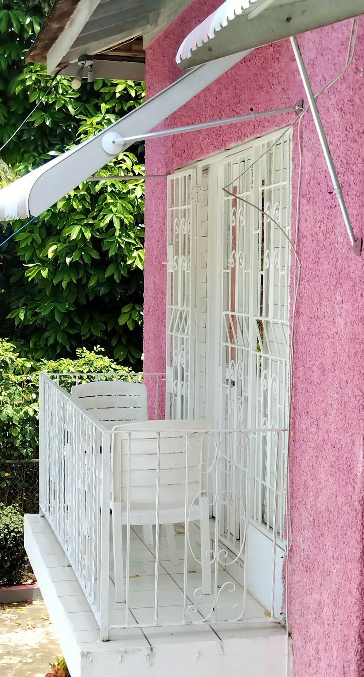 A private balcony features a white metal railing and a small white table with chairs. Sunlight reflects off the balcony surface, which is part of a pink exterior wall decorated with white window bars. Lush green foliage is visible in the background.